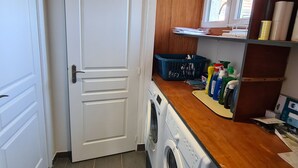 A laundry room with a washing machine, a dryer, a blue laundry basket, and shelves with cleaning supplies.