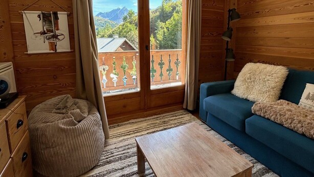 Une pièce chaleureuse aux murs en bois, avec un canapé bleu, une table basse en bois et une vue sur un balcon doté d’une balustrade en bois et de plantes vertes.