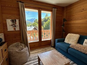 Une pièce chaleureuse aux murs en bois, avec un canapé bleu, une table basse en bois et une vue sur un balcon doté d’une balustrade en bois et de plantes vertes.