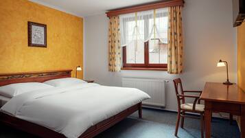 Habitación de hotel con cama de madera, un escritorio, una silla, ventana con cortinas, radiador y un cuadro en la pared.
