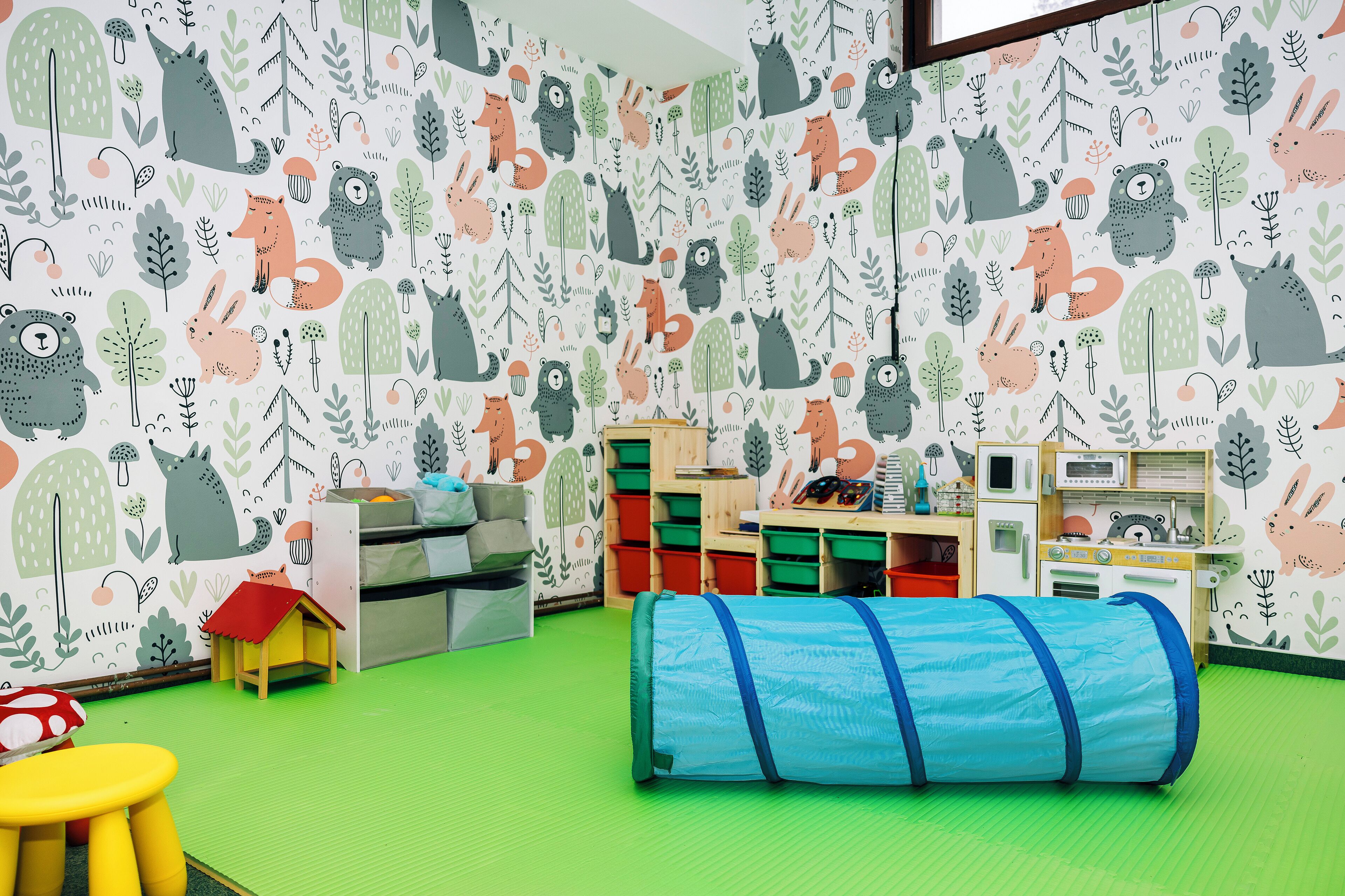 A children's playroom with a green floor, a blue tunnel, and a wall decorated with animal and plant illustrations.