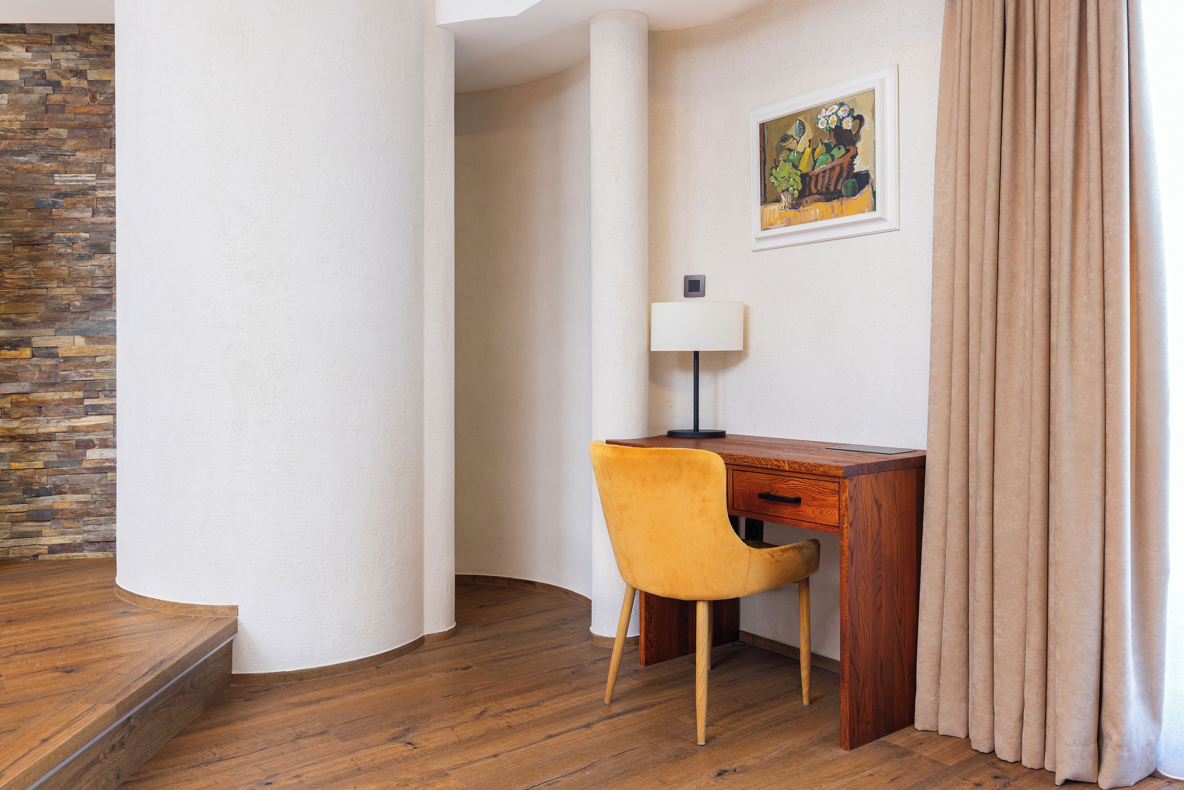 A room with a wooden desk, a yellow chair, a lamp, and a painting on the wall.