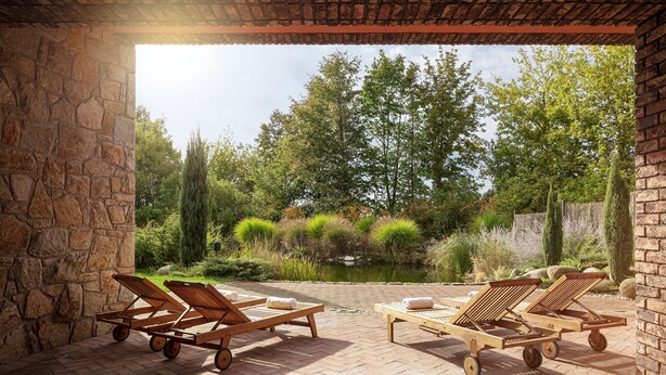 Un patio con dos sillones de madera y una mesa, un muro de piedra, y vistas a un estanque y árboles.