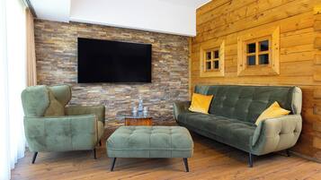 Ein gemütliches Wohnzimmer mit Steinwand, Holzvertäfelung, einem Flachbildfernseher, einem grünen Sofa mit gelben Kissen und einem passenden Hocker.