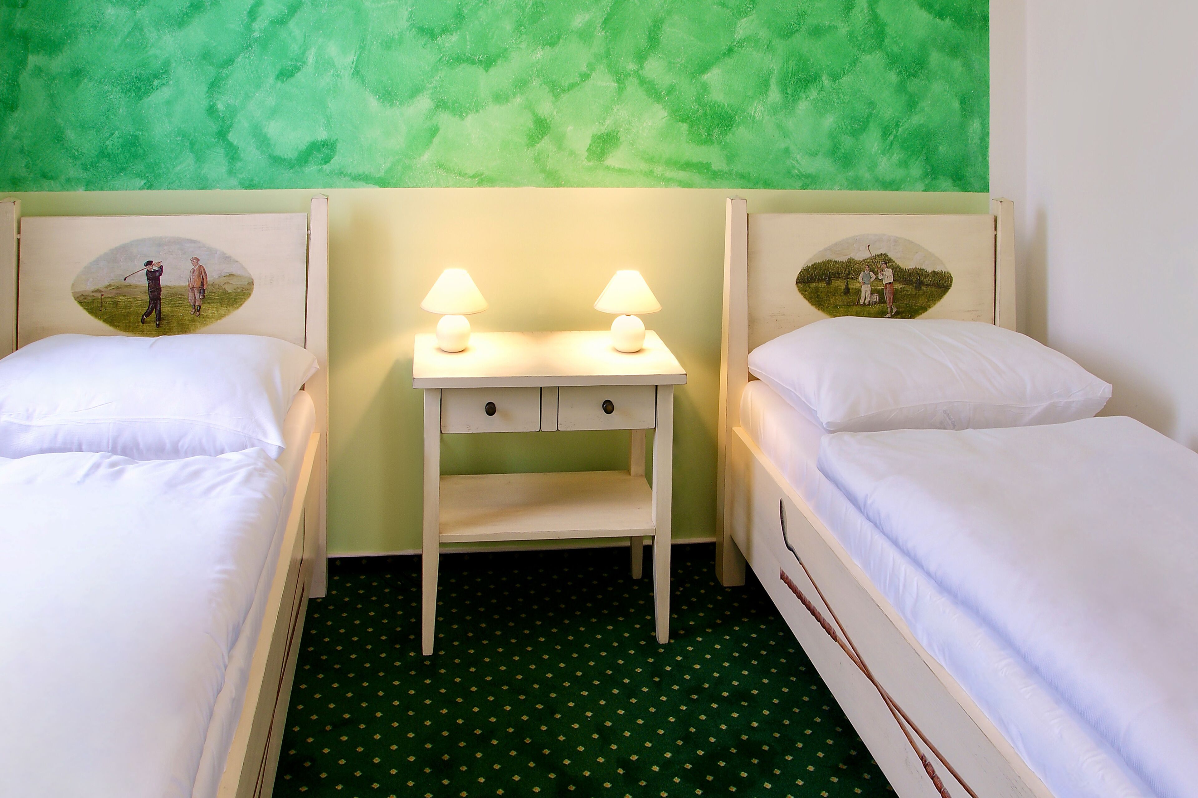 Quarto com duas camas, uma mesinha de cabeceira e luminárias, e um tapete verde estampado.
