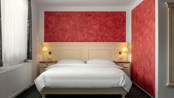 Una cama bien hecha con sábanas blancas, dos mesitas de noche con lámparas, una pared texturizada de color rojo y un radiador.