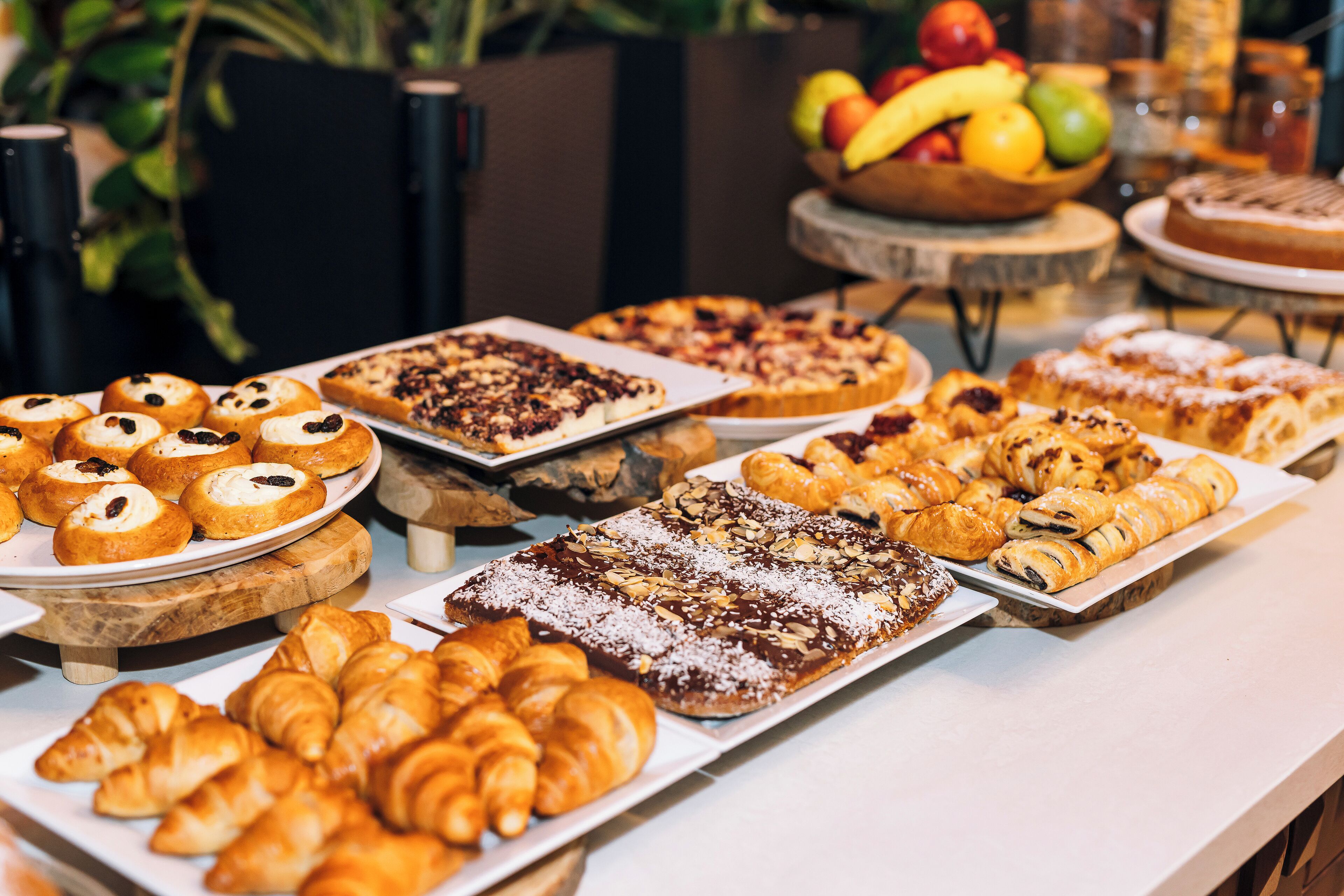 Eine Auswahl an Gebäcken und Desserts, darunter Croissants, Muffins und Kuchen, mit einer Obstschale im Hintergrund.
