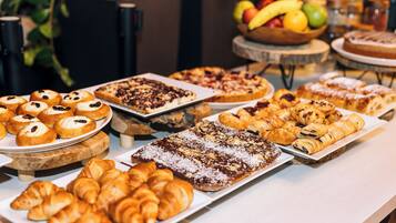 A variety of pastries and desserts on display, including croissants, muffins, and cakes, with a fruit bowl in the background.