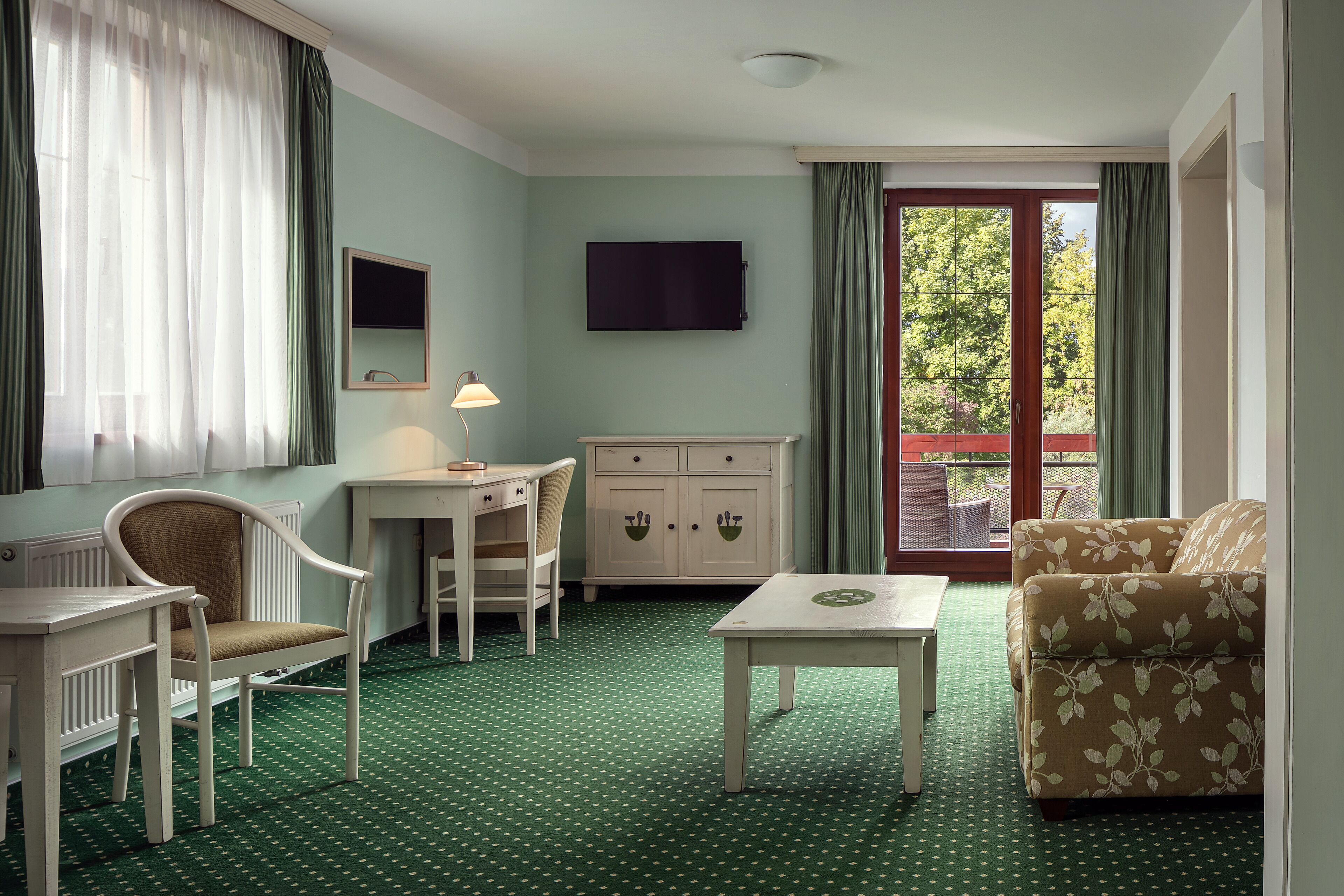 A room with a green carpet, a sofa, a chair, a table, a TV, and a window with curtains.