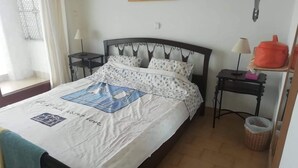 A neatly made bed with a patterned bedspread, a bedside table with a lamp, and a small shelf with a red container. - Nice House With sea View in Spetses, Greece (Spetses)
