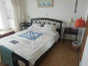 Una cama bien tendida con una colcha estampada, una mesita de noche con una lámpara y un pequeño estante con un recipiente rojo.