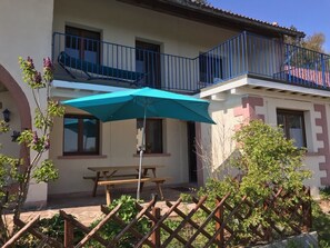 Un bâtiment à deux étages doté d’un balcon, d’un banc en bois et d’un parasol bleu.