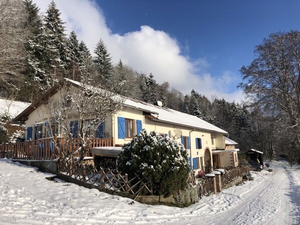 Une maison recouverte de neige, avec une clôture en bois, un arbre également recouvert de neige et un ciel dégagé.