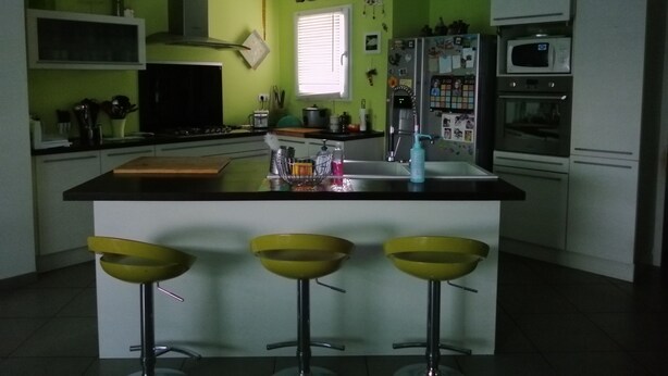 Una cocina con una isla central, tres taburetes amarillos, un refrigerador y un microondas.