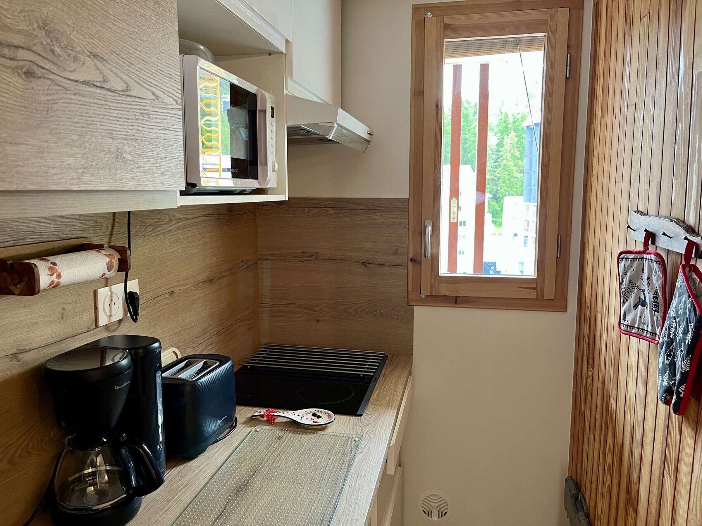 A compact kitchen with a microwave, toaster, and coffee maker. A window with vertical blinds provides natural light.