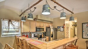 Une cuisine avec des meubles de rangement en bois, un îlot central doté de tabourets de bar, un luminaire suspendu et une fenêtre avec des stores.