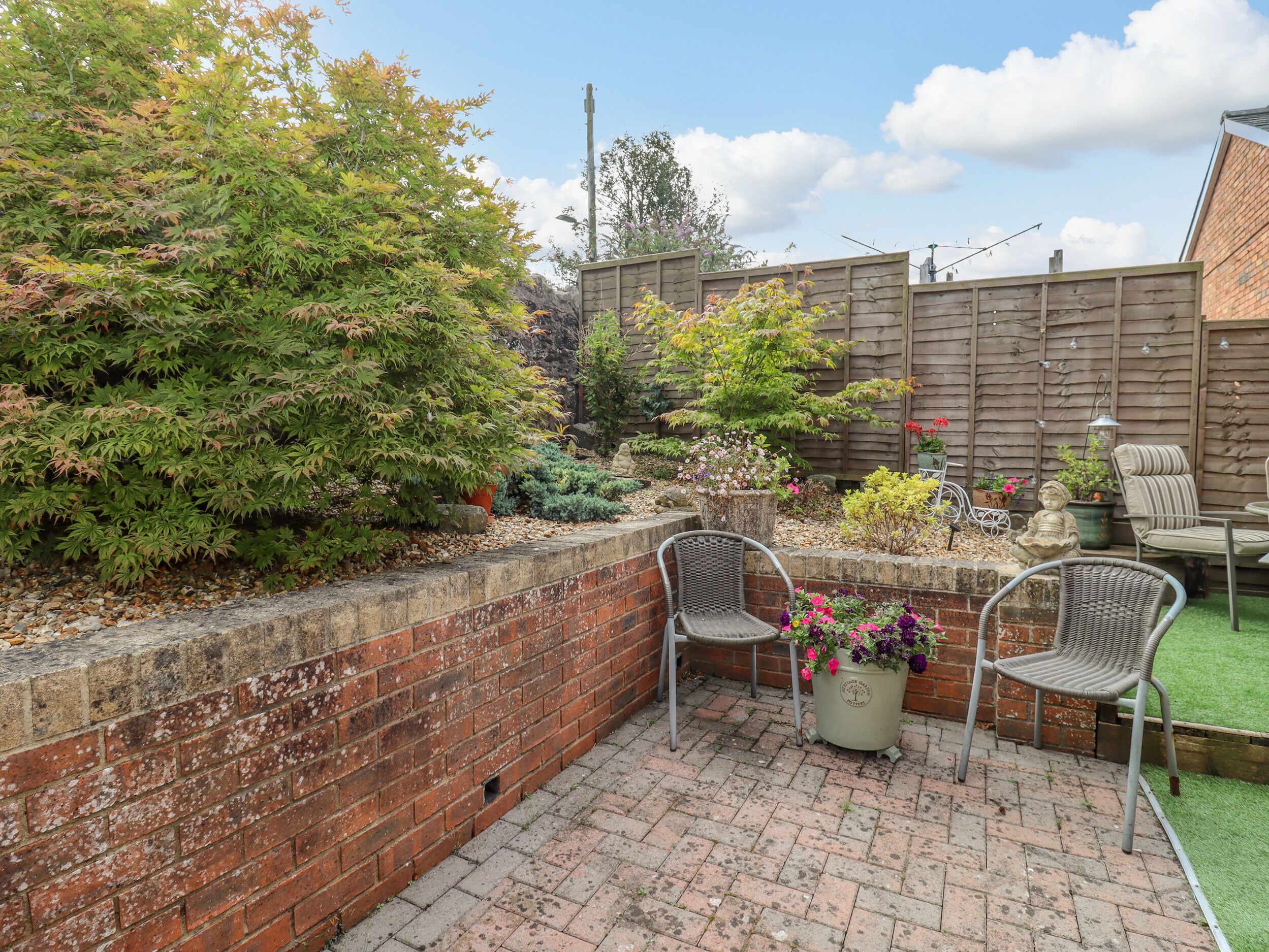 A patio area with brick flooring, two wicker chairs, a potted plant with flowers, and a wooden fence in the background.