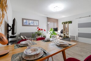 Un coin repas avec une table en bois dressée pour quatre personnes, entouré d’un salon moderne comprenant un canapé gris, un téléviseur et des œuvres d’art accrochées au mur.