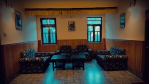 A room with wooden paneled walls, a herringbone floor, and floral-patterned sofas and chairs. There are two windows with curtains, a table, and a painting on the wall. - Rufina Norling Zimkhang (Chungthang)