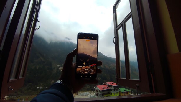 A person holding a smartphone with a camera app open, displaying a view of a mountainous landscape through a window. - Rufina Norling Zimkhang (Chungthang)