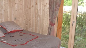 Makuuhuoneessa on puinen seinä, harmaalla tekstiilillä päällystetty sänky, jossa on punaiset koristeet, ja ikkunasta avautuu näkymä vehreään maisemaan.