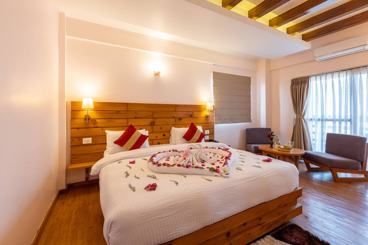 A modern hotel room with a wooden headboard, a neatly made bed with a towel and flowers, a small round table with a vase, and a comfortable chair.
