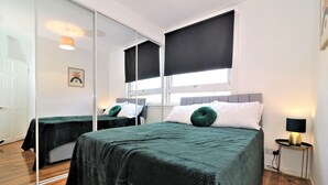 Ein Hotelzimmer mit zwei Betten, einem großen Spiegel, einem Fenster mit Verdunkelungsvorhang und einem Nachttisch mit Lampe.