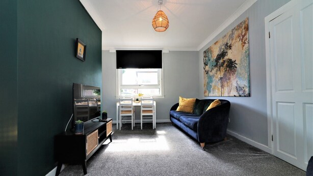 Ein kompakter Wohnraum mit einer dunkelgrünen Wand, einer schwarzen Unterhaltungsecke, einem blauen Sofa mit gelben Kissen, einem weißen Tisch mit Stühlen und einem großen abstrakten Gemälde.