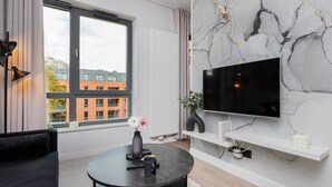 Ein modernes Wohnzimmer mit großem Fenster, einem wandmontierten Flachbildfernseher, einem runden schwarzen Tisch mit Vase und Fernbedienung sowie einer schwarzen Couch.