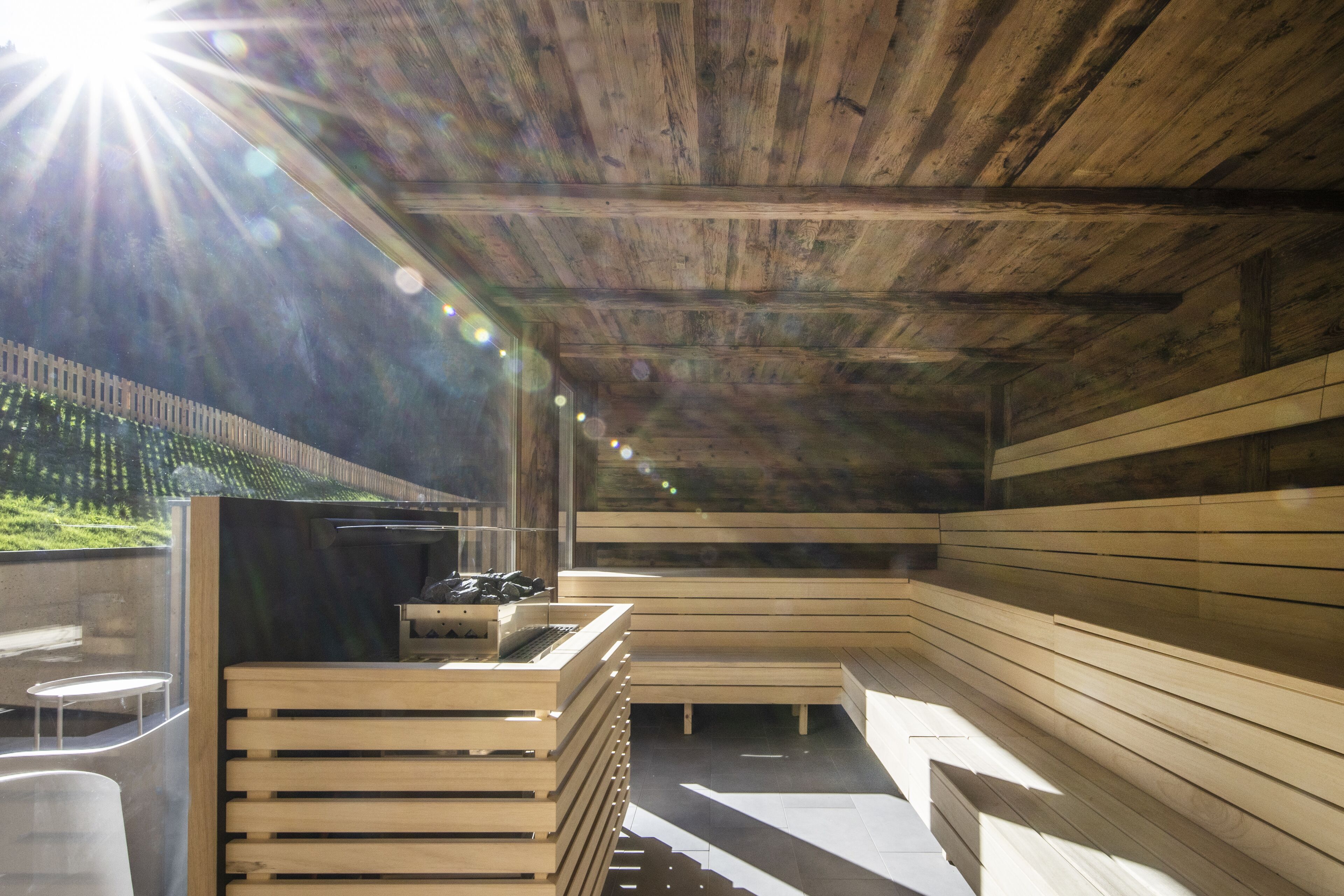 A sauna with wooden benches and a bench area, a small table with items, and a view of greenery outside.