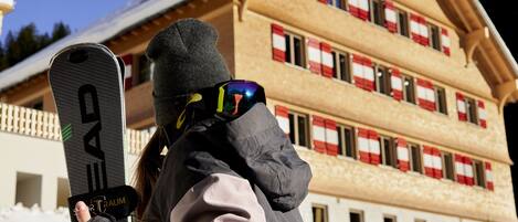 Una persona sosteniendo una tabla de snowboard con la marca "HEAD" a la vista, de pie frente a un edificio con techo inclinado y varias ventanas, algunas con cortinas a rayas rojas y blancas.