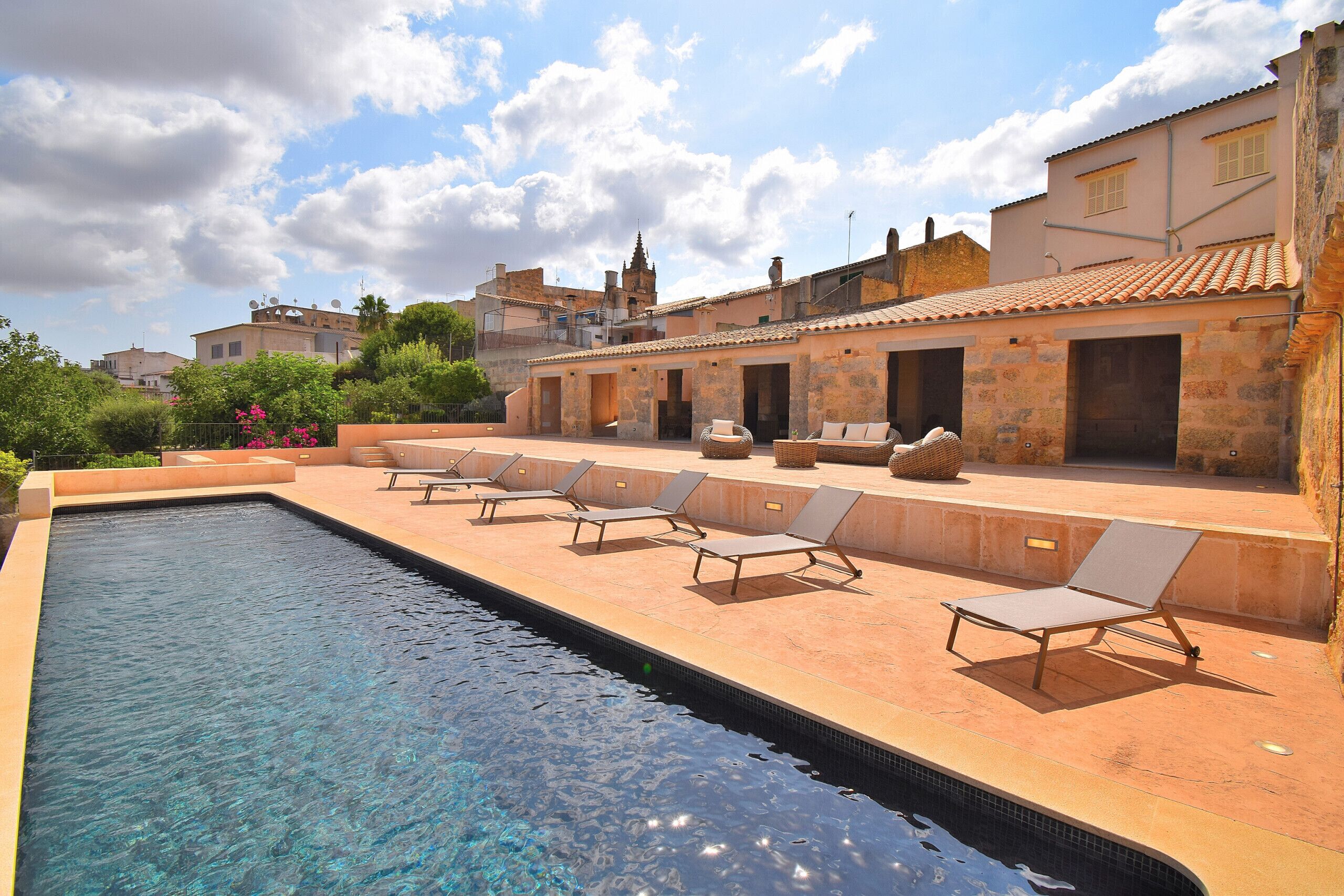 A swimming pool with lounge chairs, a stone building with a tiled roof, and a view of surrounding buildings.