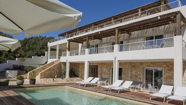 A poolside area with lounge chairs and umbrellas, a stone building with a wooden deck, and a staircase leading to a balcony. - Mercury Beach Hotel (Sant'Antioco)