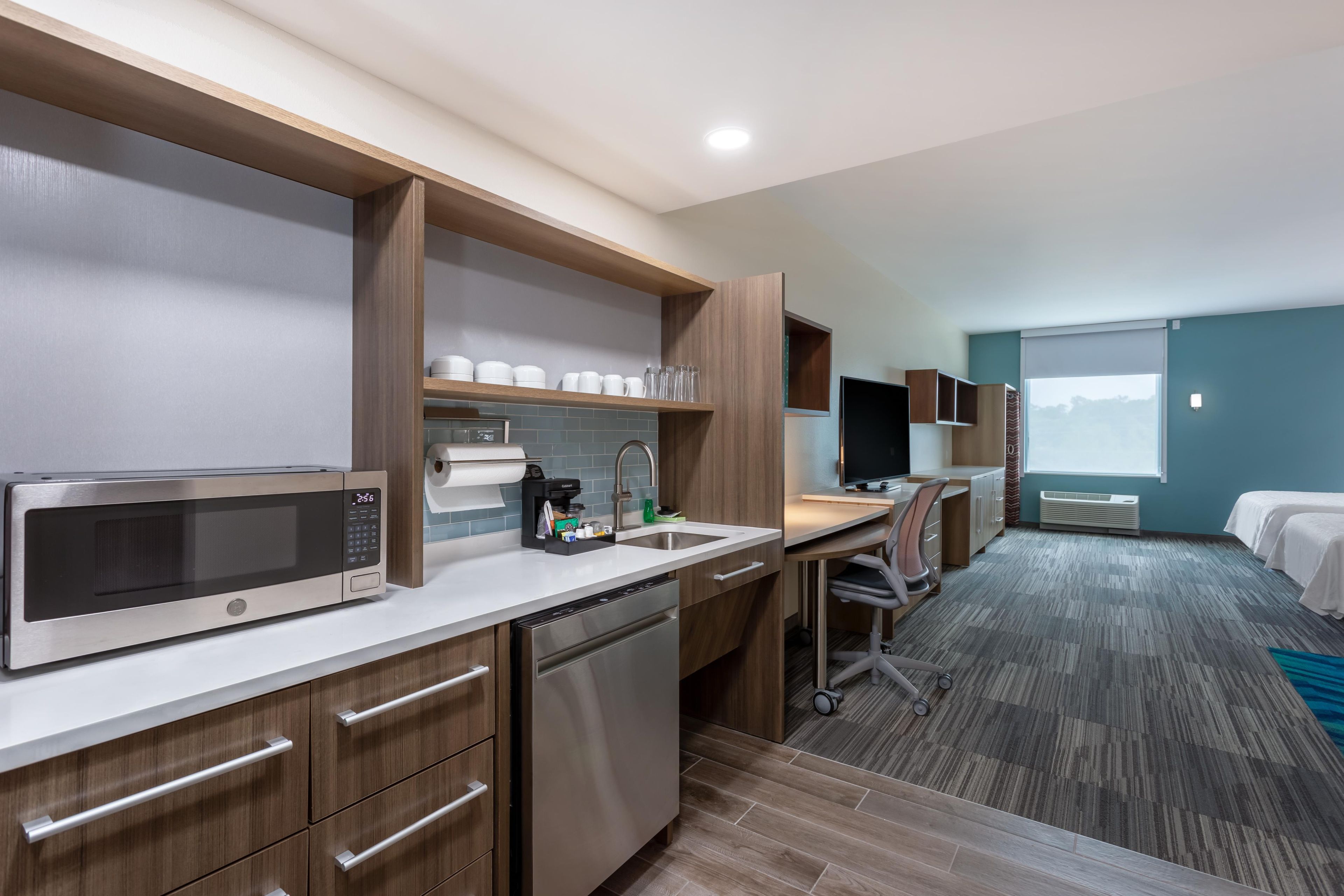 A modern hotel room with a kitchenette, a desk, a bed, and a large window.