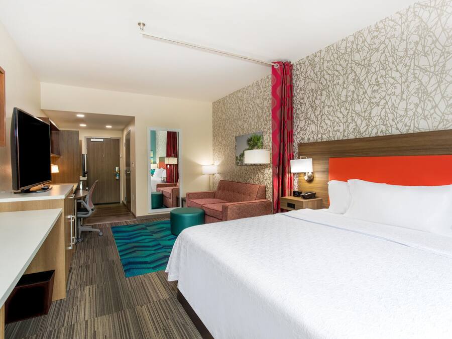 Habitación de hotel con cama, escritorio, televisor, silla, mesita de noche y baño visible a través de una puerta abierta.