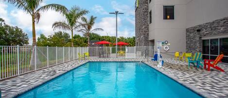 A swimming pool with a tiled deck, surrounded by a white fence, and colorful chairs and umbrellas.