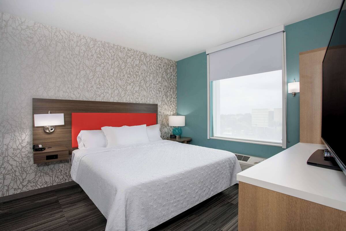 A hotel room with a bed, a desk, a TV, a window, and a nightstand with a lamp.