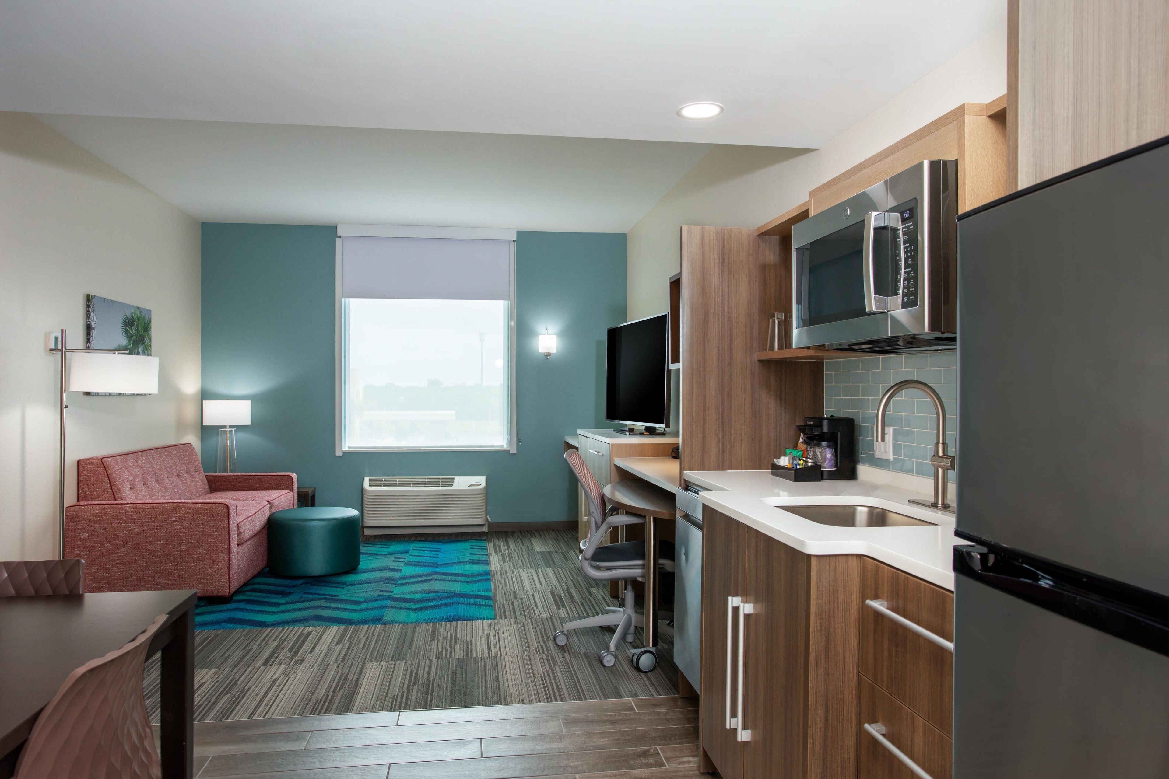 A compact hotel room with a kitchenette, a seating area with a sofa and a small table, a large window, and a view of the cityscape.