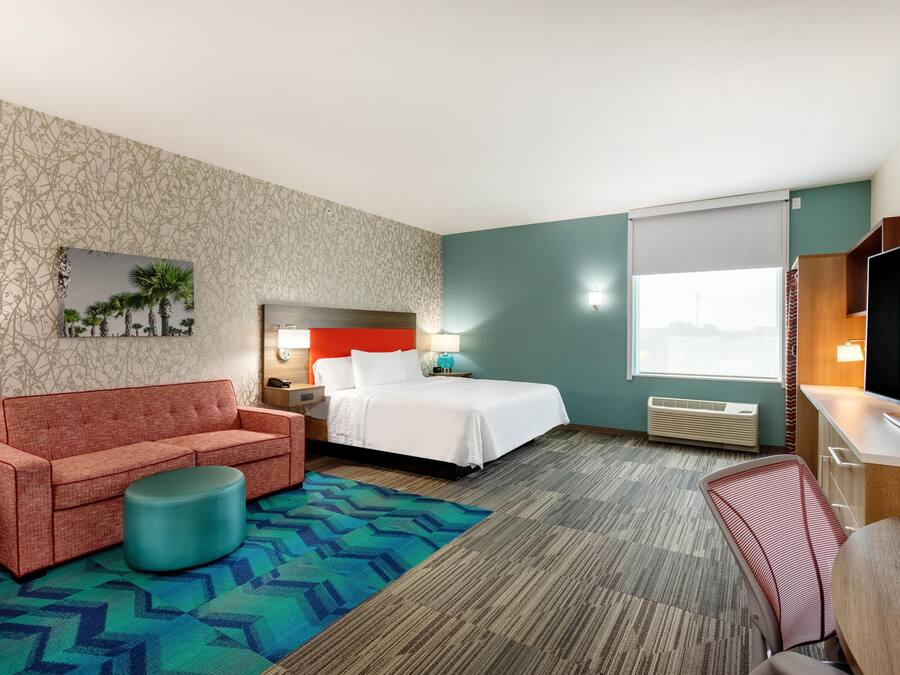 A hotel room with a bed, a sofa, a small ottoman, a desk, and a television.