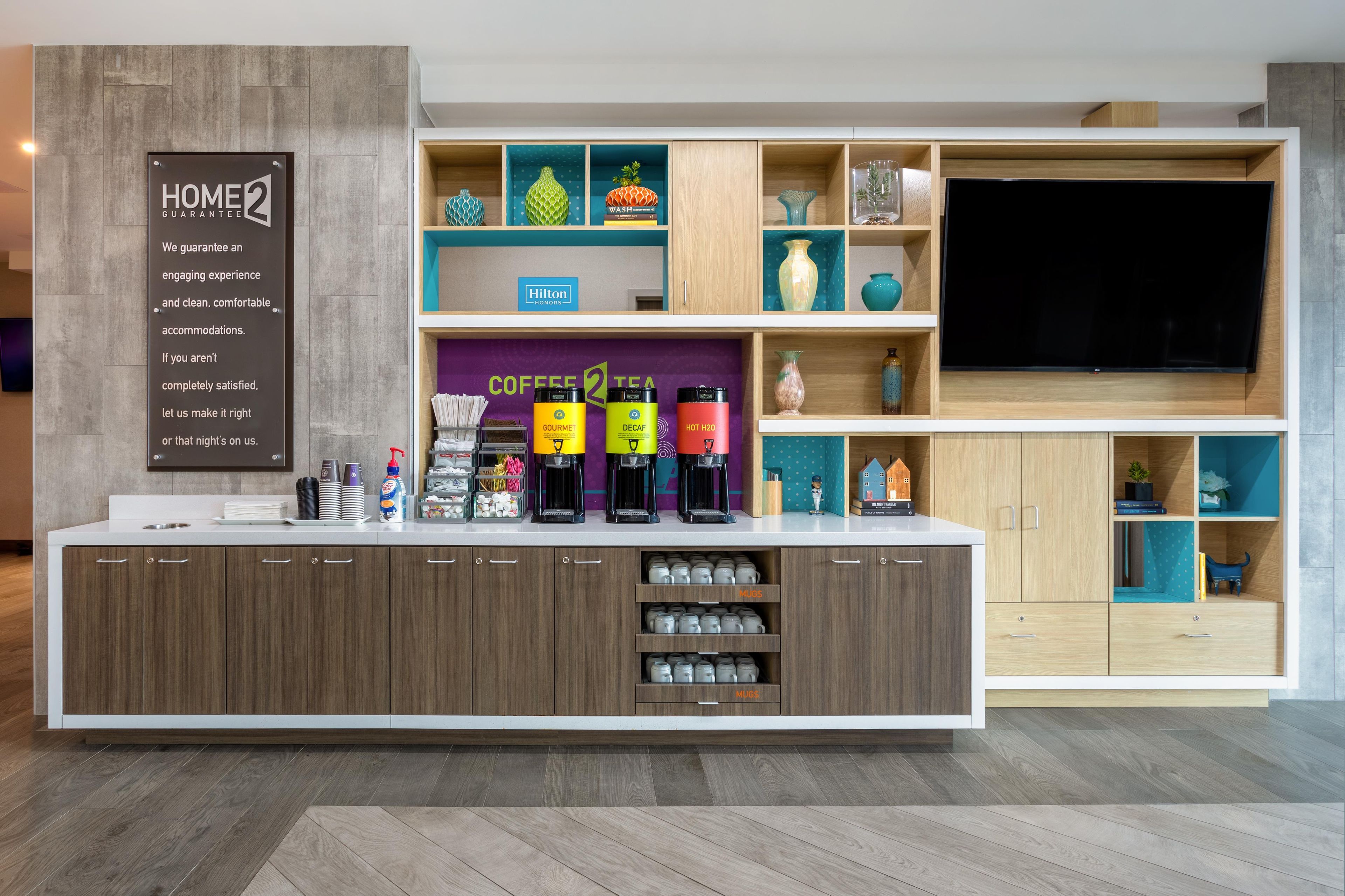 A modern hotel lobby with a coffee station, a large flat-screen TV, and a sign for the hotel's guarantee.