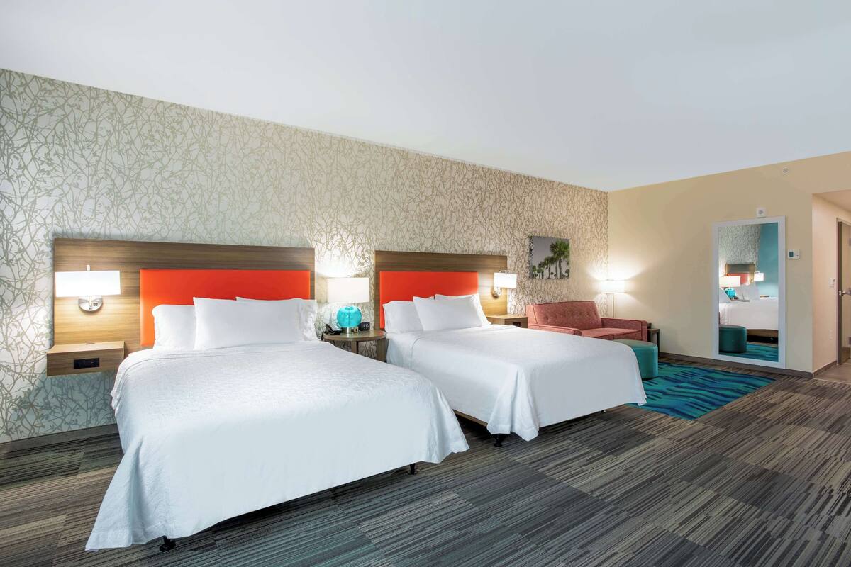 A hotel room with two beds, a red accent wall, and a large mirror.