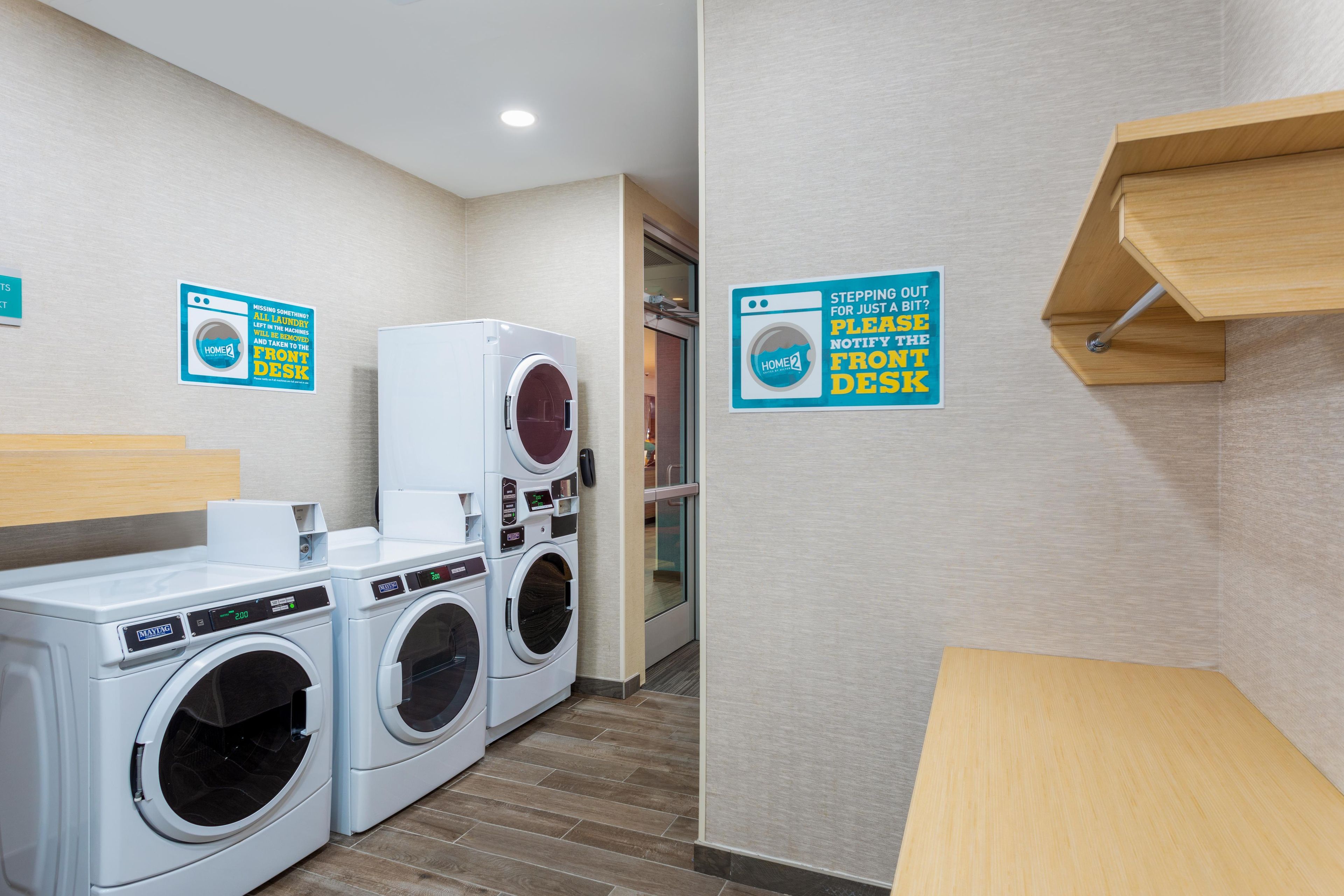 A laundry room with multiple washing machines, a drying rack, and signs instructing to notify the front desk if stepping out for a bit.