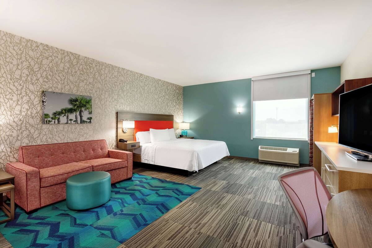 A hotel room with a bed, a red sofa, a small round ottoman, a desk with a TV, and a window with a view.