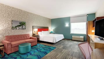 Habitación de hotel con cama, sofá rojo, reposapiés redondo pequeño, escritorio con televisor y ventana con vistas.