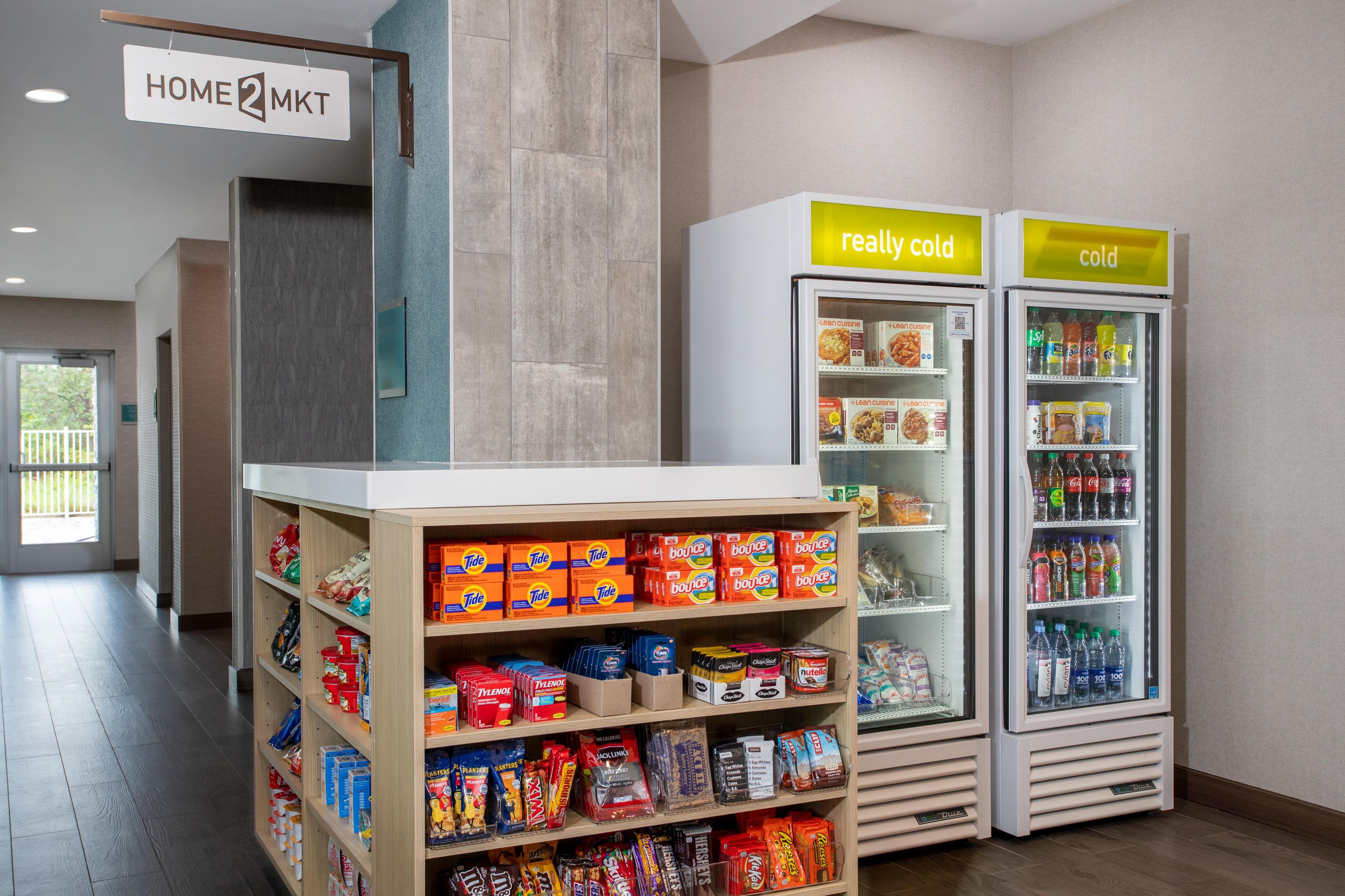 A hotel lobby with a self-service kiosk, refrigerated beverage dispensers, and shelves stocked with snacks and household items.