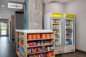 A hotel lobby with a self-service kiosk, refrigerated beverage dispensers, and shelves stocked with snacks and household items. - Home2 Suites by Hilton Vero Beach I-95 (Vero Beach)