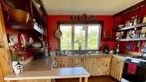 A kitchen with wooden cabinets, a central island with stools, a stove, and a window with a view of greenery. - Spacious home w/riverview, minutes from state parks and ocean beaches, sleeps 11 (East Machias)