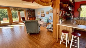 A spacious living room with a wooden floor, a dining area with a table and chairs, and a kitchen with wooden cabinets and a window. - Spacious home w/riverview, minutes from state parks and ocean beaches, sleeps 11 (East Machias)