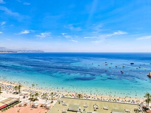 Une plage aux eaux d’un bleu limpide, où l’on peut voir de nombreux bateaux, et une zone de plage très fréquentée, parsemée de parasols et de gens.
