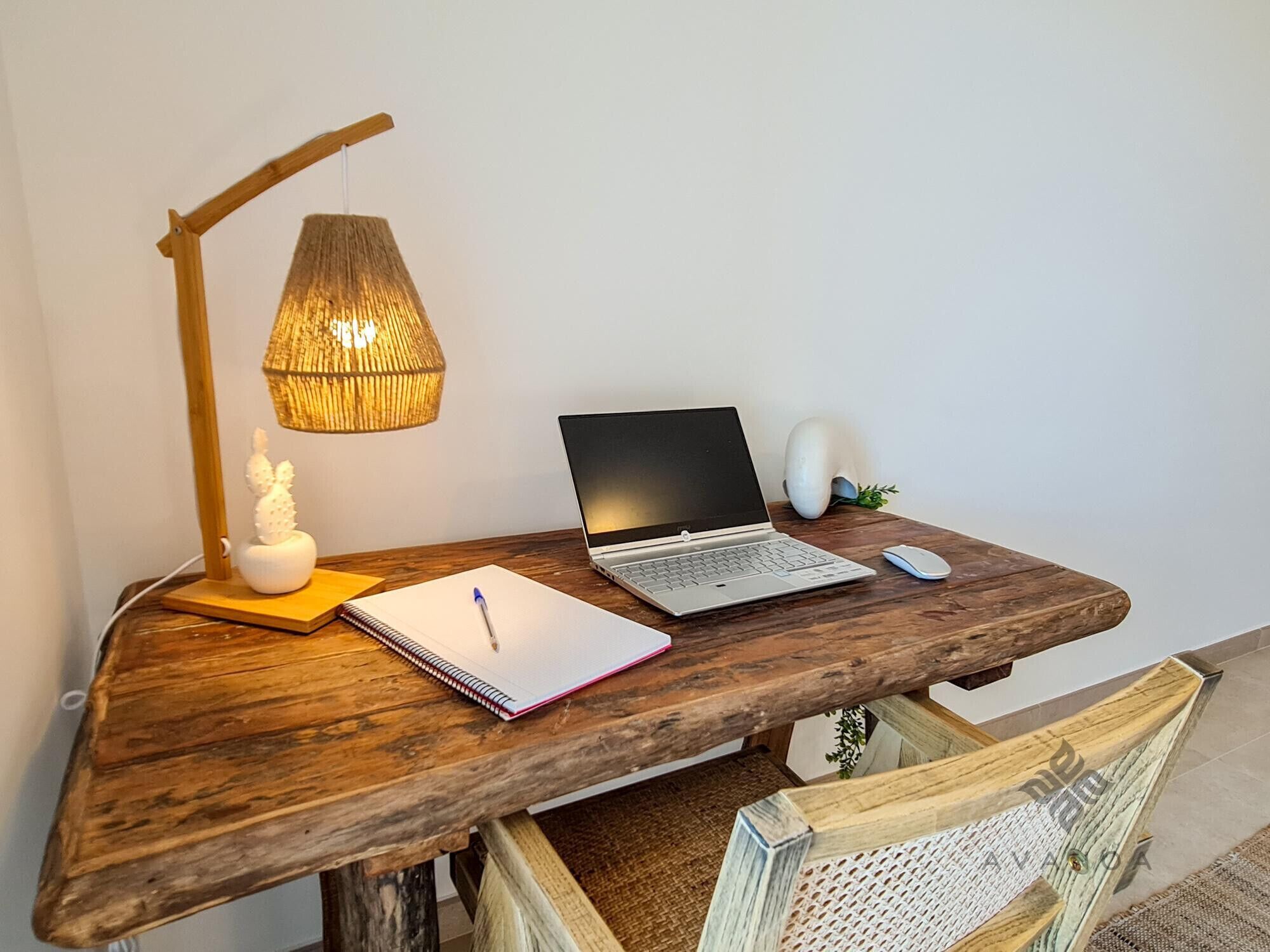 A wooden desk with a laptop, a notebook, a pen, a mouse, a lamp, and a decorative item.
